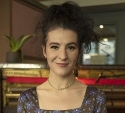Color bio photo of Jesse Paris Smith standing in front of a piano wearing dark flowered dress and necklace smiling into camera.