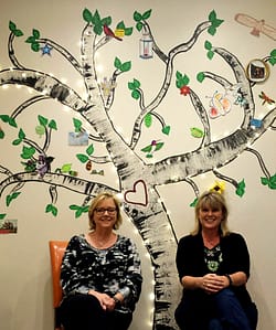Amber and Julee sitting in front of the memory tree on the wall of their space.