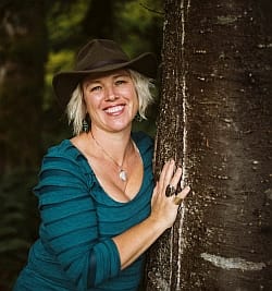 Bio photo of Kylee wearing blue shirt and dark hat, leaning against a tree, smiling into camera
